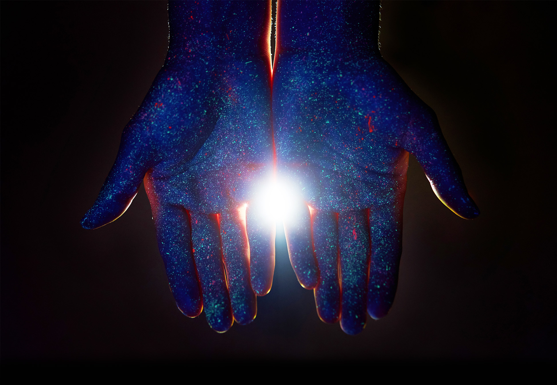 A pair of open hands, palms facing and pointing downwards, painted in blue ultraviolet paint with a light shining through from behind.